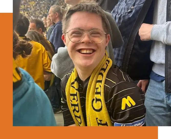 Zum Tivoli begleiteter Junge mit Down-Syndrom und Fußball Fan- Schal sitzt im Zuschauerraum eines Fußballstadion und lächelt in die Kamera.